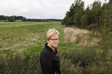 Metsäasiantuntija Marjaana Hellän takana näkyvät pelto ja Kiiskijärven rantametsät, joista ensimmäiset sikaruttoon kuolleet villisiat löytyvät. Kaakonkulmalla harva on kohdannut villisian, mutta rajan läheiset kuusikot ovat myllättyjä ja juuret paistavat. (Kuvaaja: Seppo Samuli)